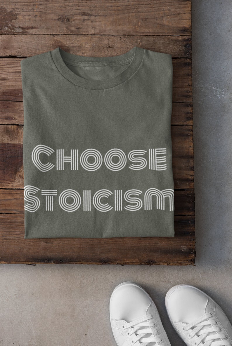 May include: A green t-shirt with the text "CHOOSE STOICISM" in white, stylised font. The shirt is folded and laid on a wooden surface. Two white trainers are on a grey surface below the shirt.