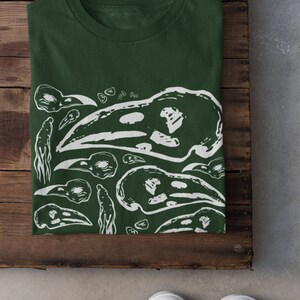 Op de afbeelding: Donkergroen t-shirt met een witte grafische print van vogel schedels en abstracte vormen. Het shirt is opgevouwen op een houten oppervlak, met witte sneakers zichtbaar. Het ontwerp wordt herhaald.