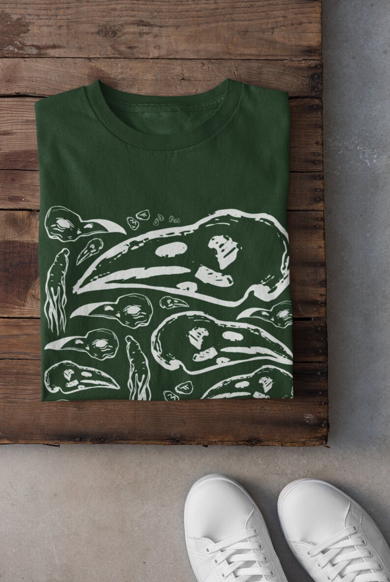 Op de afbeelding: Donkergroen t-shirt met een witte grafische print van vogel schedels en abstracte vormen. Het shirt is opgevouwen op een houten oppervlak, met witte sneakers zichtbaar. Het ontwerp wordt herhaald.