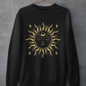 May include: Black sweatshirt with a gold sun and moon design. The sun has a face with closed eyes and a crescent moon on its forehead. There are small stars around the sun.