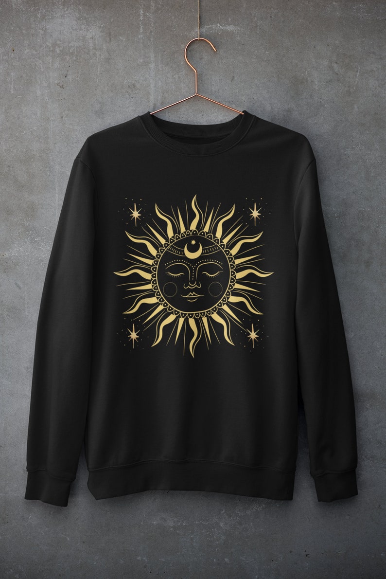 May include: Black sweatshirt with a gold sun and moon design. The sun has a face with closed eyes and a crescent moon on its forehead. There are small stars around the sun.