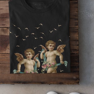 May include: Black t-shirt with a graphic print of two cherubs with wings and floral garlands, flying against a background of white birds.