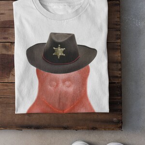May include: A white t-shirt with a red, cartoon-like figure wearing a brown cowboy hat with a gold sheriff's star. The figure has a red, textured surface.