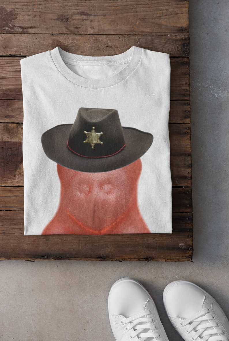 May include: A white t-shirt with a red, cartoon-like figure wearing a brown cowboy hat with a gold sheriff's star. The figure has a red, textured surface.