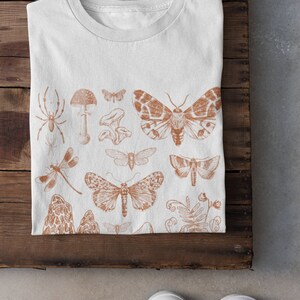 May include: White t-shirt featuring a brown print of various insects and mushrooms. The design includes butterflies, moths, a spider, a dragonfly, and mushrooms.