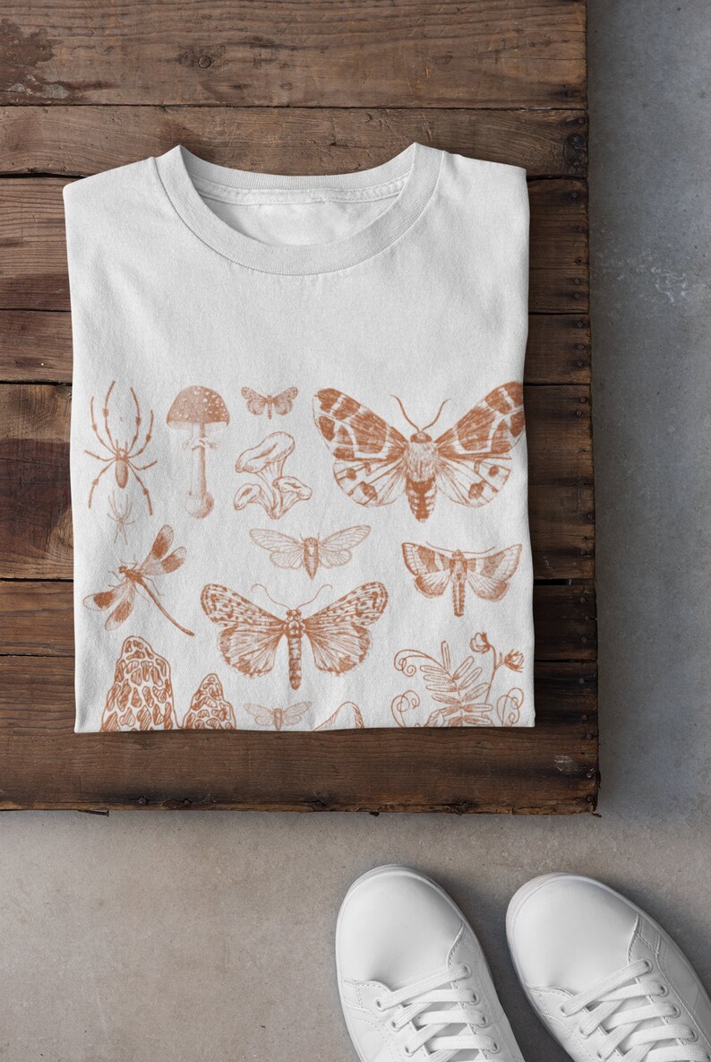May include: White t-shirt featuring a brown print of various insects and mushrooms. The design includes butterflies, moths, a spider, a dragonfly, and mushrooms.