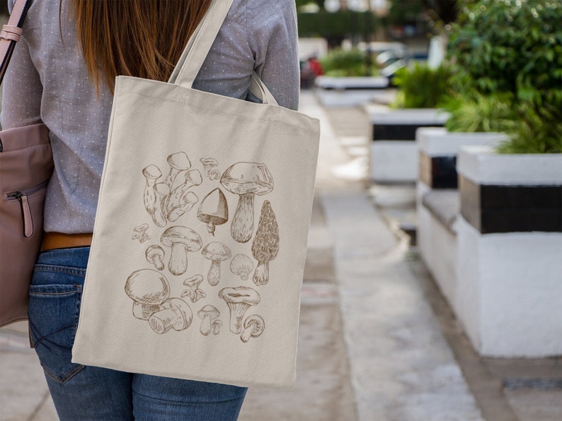 Peut inclure: Un sac fourre-tout en toile beige avec une illustration brune de diff&eacute;rents champignons. Le sac a une anse brune et est port&eacute; par une personne.