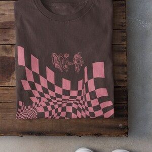 May include: A brown t-shirt featuring a pink and black checkered pattern with a graphic of three angels holding hands. The pattern is distorted to create a warped effect.