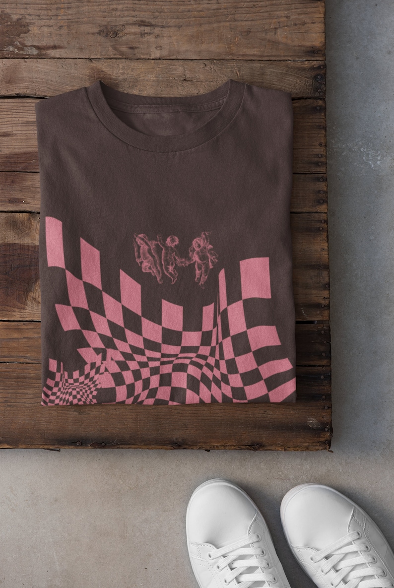 May include: A brown t-shirt featuring a pink and black checkered pattern with a graphic of three angels holding hands. The pattern is distorted to create a warped effect.