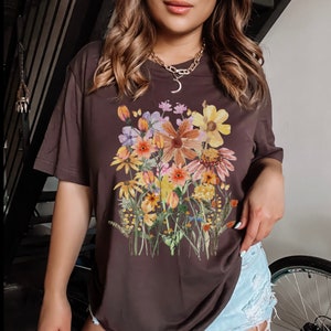 May include: A dark brown t-shirt with a watercolor floral print featuring a variety of flowers in shades of orange, yellow, pink, and purple.