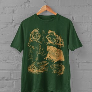 Pode incluir: Uma camiseta verde escura com uma ilustra&ccedil;&atilde;o vintage de v&aacute;rios sapos e r&atilde;s em uma cor marrom claro. O design apresenta uma variedade de poses de sapos, incluindo sentados, pulando e nadando.