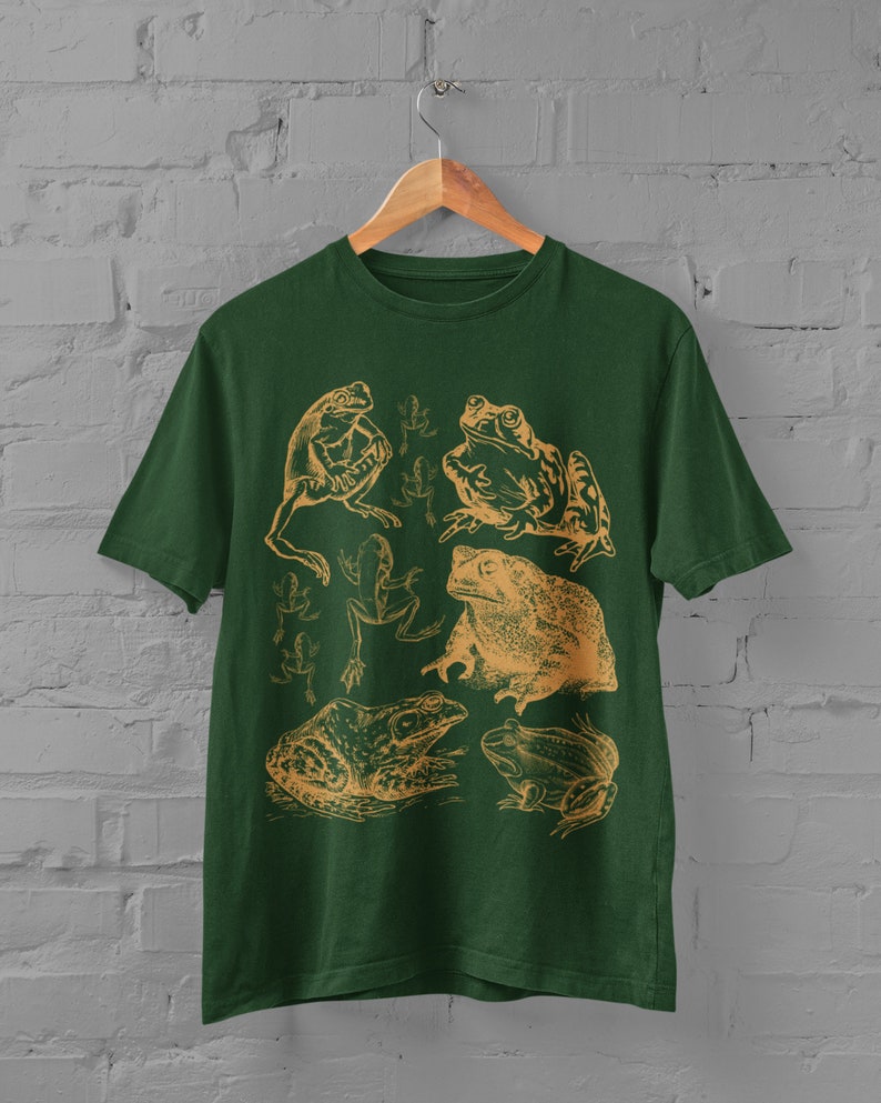 Pode incluir: Uma camiseta verde escura com uma ilustra&ccedil;&atilde;o vintage de v&aacute;rios sapos e r&atilde;s em uma cor marrom claro. O design apresenta uma variedade de poses de sapos, incluindo sentados, pulando e nadando.
