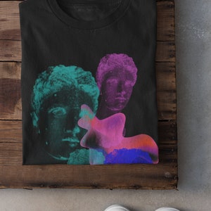 May include: Black t-shirt with a graphic print of three faces in shades of green, pink, and blue. The faces are stylized and appear to be from ancient sculptures.