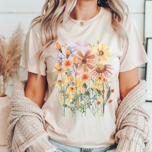 May include: A beige t-shirt with a watercolor floral print featuring yellow, orange, pink, and purple flowers. The flowers are arranged in a bouquet-like design.