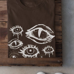 May include: A brown t-shirt featuring a white eye design. The design includes multiple stylized eyes with various shapes and sizes. The shirt is folded and placed on a wooden surface, with white sneakers in the foreground.