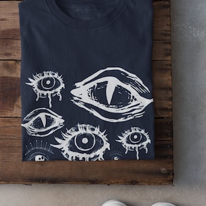 May include: A navy blue t-shirt with a white print of multiple stylized eyes. The eyes are drawn in a graphic style with dripping lines.