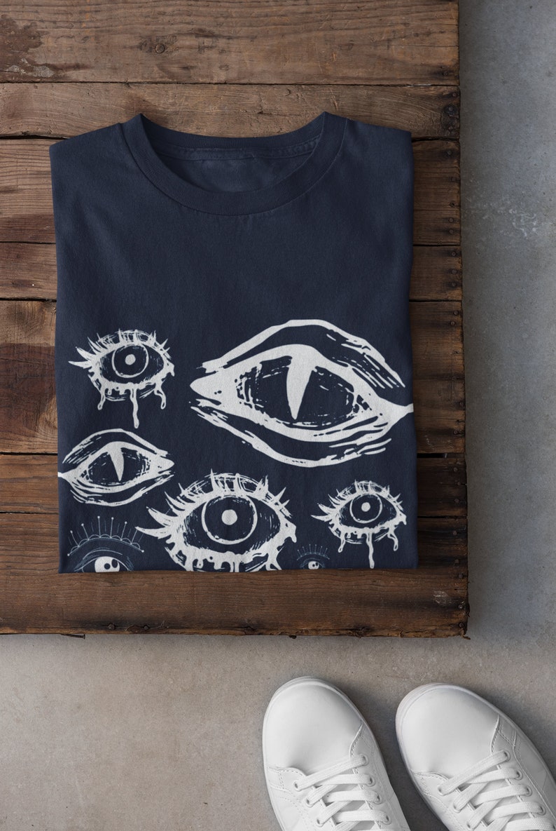 May include: A navy blue t-shirt with a white print of multiple stylized eyes. The eyes are drawn in a graphic style with dripping lines.