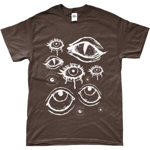 May include: A brown t-shirt with a white graphic print of multiple stylized eyes. The eyes are drawn in a variety of shapes and sizes, some with eyelashes and some with tears.
