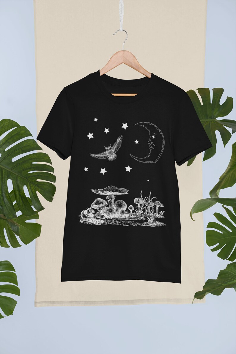May include: Black t-shirt with a white graphic design of a crescent moon, an owl flying, and mushrooms. The design is reminiscent of a night sky with stars.