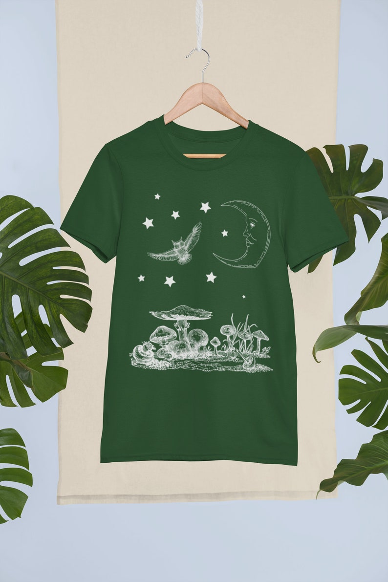 May include: A dark green t-shirt with a white graphic of a crescent moon, a flying owl, and mushrooms. The graphic is in a vintage style.