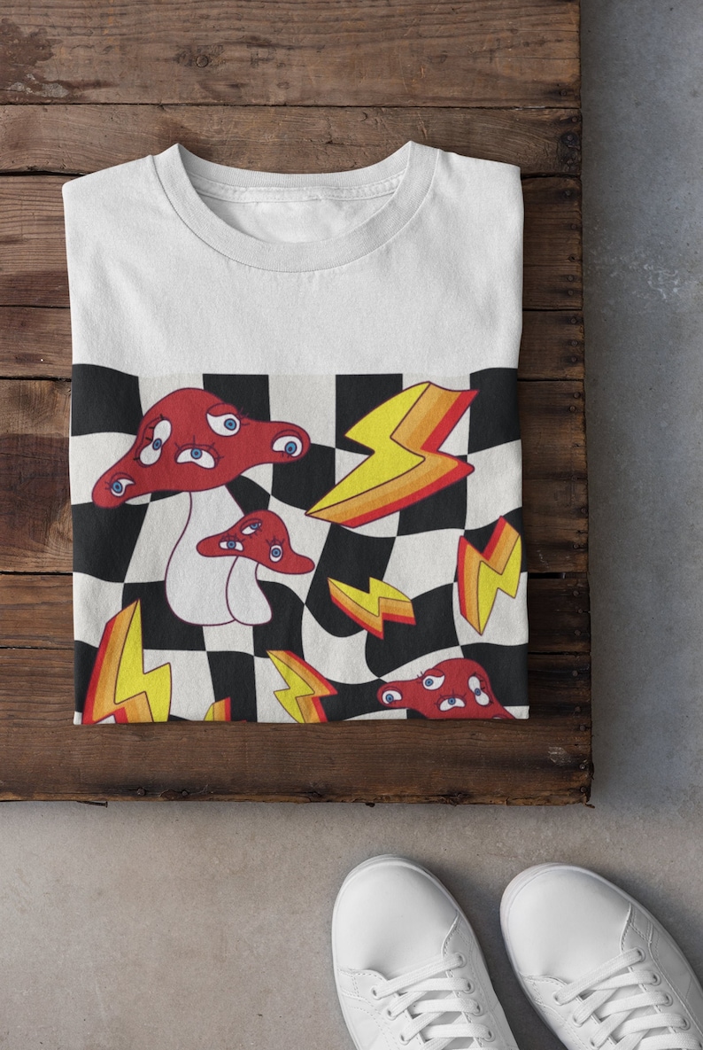 May include: White t-shirt with a checkered pattern featuring red mushrooms with multiple eyes and yellow lightning bolts.