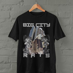 Op de afbeelding: Zwart t-shirt met de witte tekst "BIG CITY RATS". De afbeelding toont drie ratten voor een skyline met rookeffecten. Het ontwerp is gedrukt op een zwart t-shirt.
