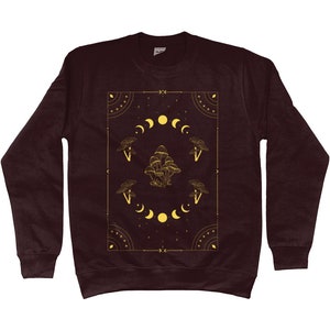 May include: A maroon sweatshirt with a gold graphic design featuring mushrooms, moons, and stars. The design is framed by a geometric border.