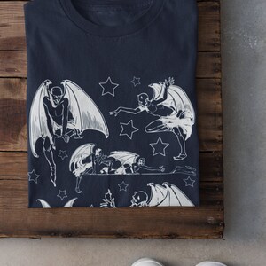 May include: A navy blue t-shirt with a white print of multiple winged figures and stars. The figures are in various poses, some with wings spread and some with wings folded. The stars are scattered around the figures.