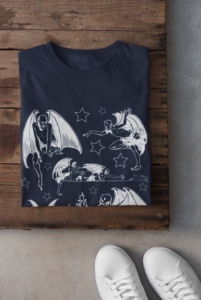 May include: A navy blue t-shirt with a white print of multiple winged figures and stars. The figures are in various poses, some with wings spread and some with wings folded. The stars are scattered around the figures.