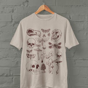 Puede incluir: Una camiseta beige con un estampado marrón de varios cráneos, insectos y setas. El estampado incluye una libélula, un cráneo con un pico de pájaro y un cráneo con una nariz larga y puntiaguda.
