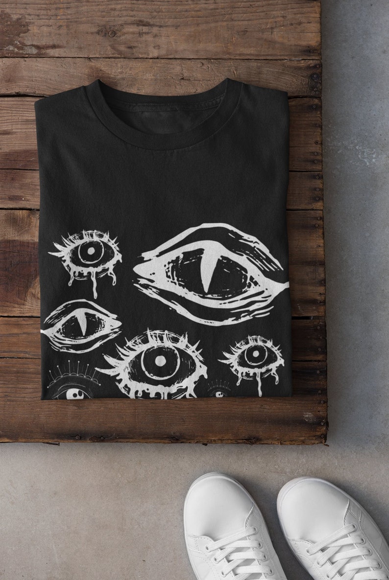 May include: Black t-shirt featuring a white eye design. The graphic includes various eye illustrations with different shapes and styles. The shirt is folded on a wooden surface, with white sneakers visible in the foreground.