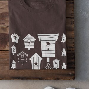 May include: A brown t-shirt featuring a white design of various birdhouses. The design includes a variety of shapes and sizes of birdhouses.