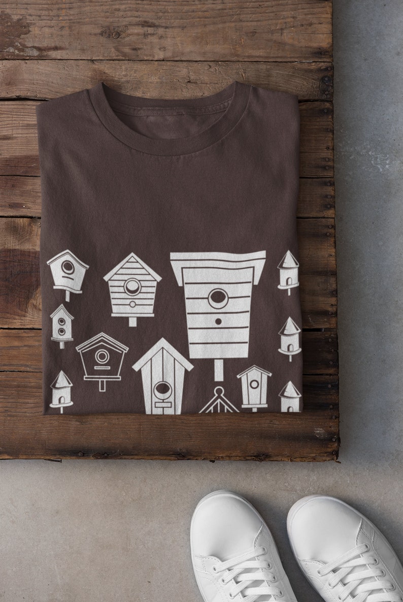 May include: A brown t-shirt featuring a white design of various birdhouses. The design includes a variety of shapes and sizes of birdhouses.