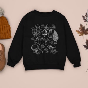 Puede incluir: Sudadera negra con un gr&aacute;fico blanco de varios champi&ntilde;ones. La sudadera est&aacute; colocada sobre una superficie marr&oacute;n claro con un gorro de punto marr&oacute;n, botas marrones y hojas de arce marrones.