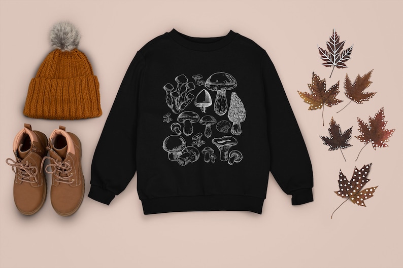 Puede incluir: Sudadera negra con un gr&aacute;fico blanco de varios champi&ntilde;ones. La sudadera est&aacute; colocada sobre una superficie marr&oacute;n claro con un gorro de punto marr&oacute;n, botas marrones y hojas de arce marrones.