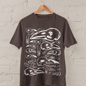 Op de afbeelding: Donkerbruin t-shirt met een witte grafische print van verschillende vogel schedels. Het ontwerp toont schedels in verschillende maten en ori&euml;ntaties, waardoor een herhalend patroon ontstaat. Het t-shirt hangt aan een houten hanger voor een witte bakstenen muur.