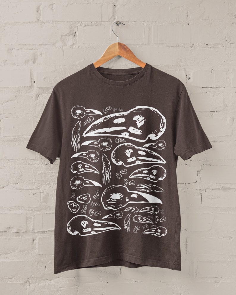 Op de afbeelding: Donkerbruin t-shirt met een witte grafische print van verschillende vogel schedels. Het ontwerp toont schedels in verschillende maten en ori&euml;ntaties, waardoor een herhalend patroon ontstaat. Het t-shirt hangt aan een houten hanger voor een witte bakstenen muur.