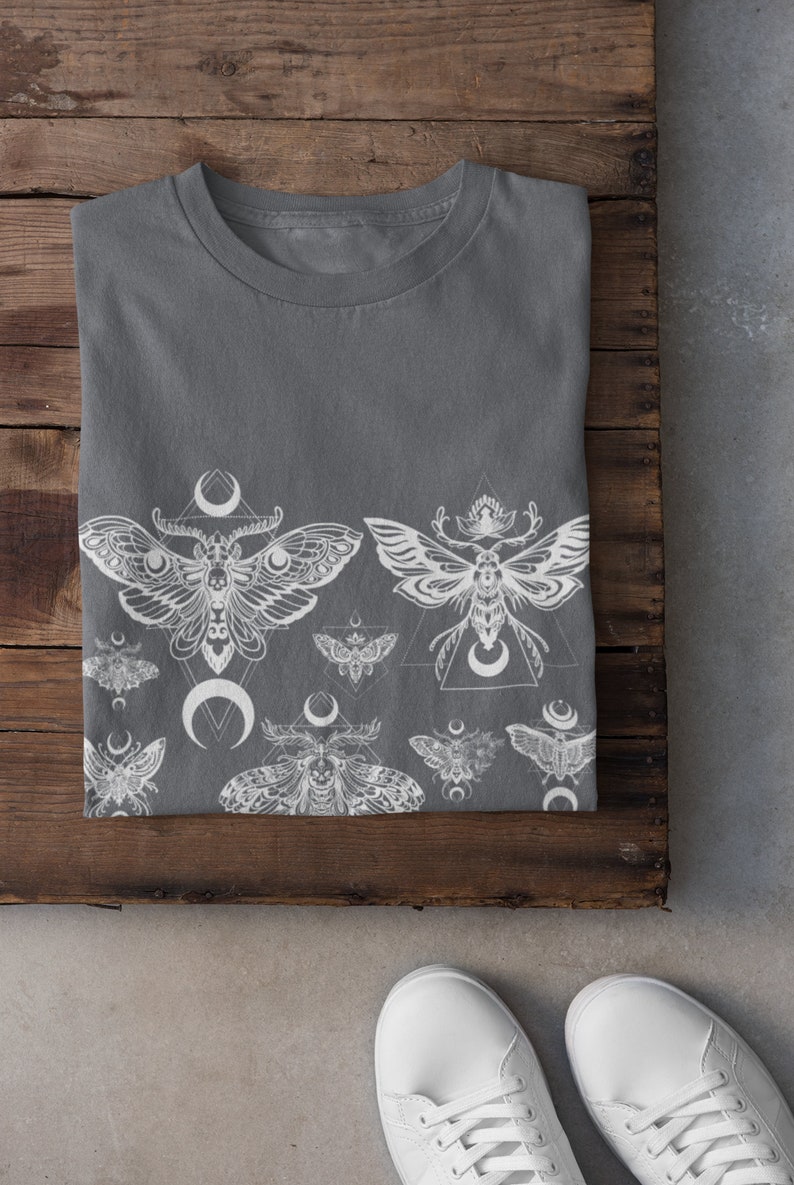 May include: A gray t-shirt with a white print of moths and crescent moons. The moths are stylized and have intricate details. The print is centered on the shirt and extends from the bottom hem to the middle of the shirt.