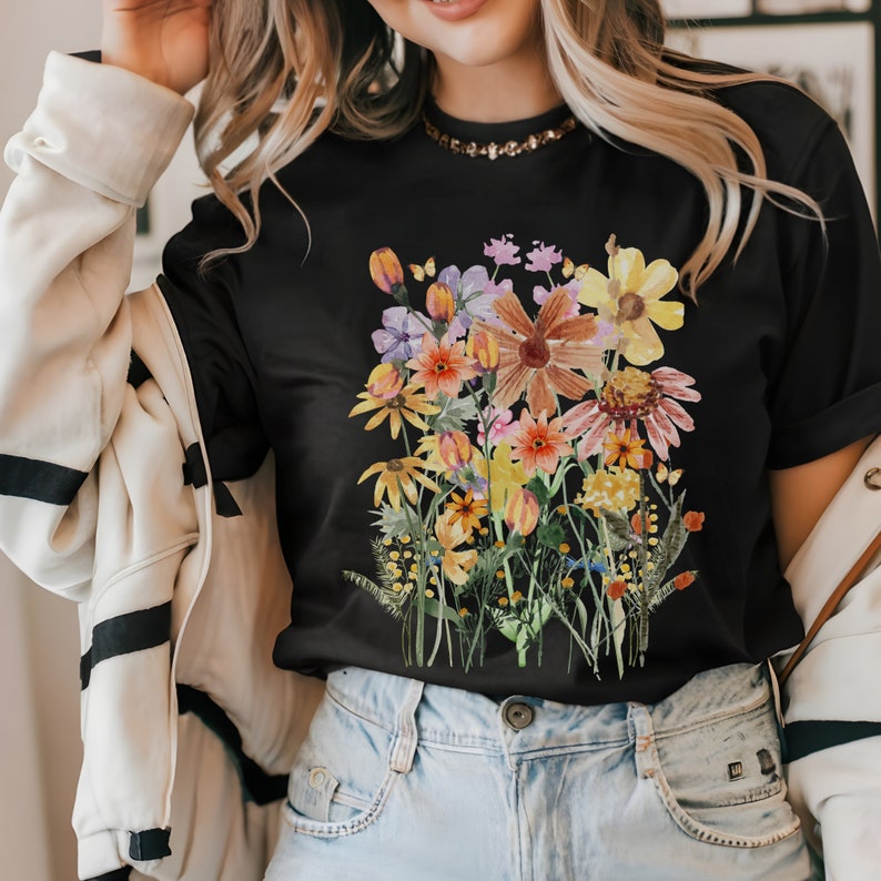 May include: A black t-shirt with a colorful floral print. The print features a variety of flowers in shades of pink, orange, yellow, and purple. The flowers are arranged in a bouquet-like shape and are surrounded by green leaves.
