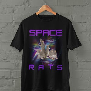 Puede incluir: Camiseta negra con las palabras "SPACE RATS" en letras moradas. El diseño incluye un gráfico de varias ratas grises sobre un fondo abstracto y colorido con tonos morados, azules y blancos.