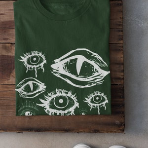 May include: A dark green t-shirt with a white print of multiple stylized eyes. The eyes are drawn in a creepy, abstract style.