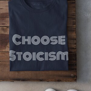 May include: A navy blue t-shirt with the text "CHOOSE STOICISM" printed in white. The text is in a retro, stylized font.