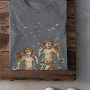 May include: A gray t-shirt with a print of two cherubs with wings and flowers. The cherubs are holding blue ribbons. There are white birds flying in the background.
