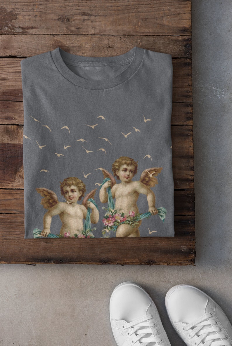 May include: A gray t-shirt with a print of two cherubs with wings and flowers. The cherubs are holding blue ribbons. There are white birds flying in the background.