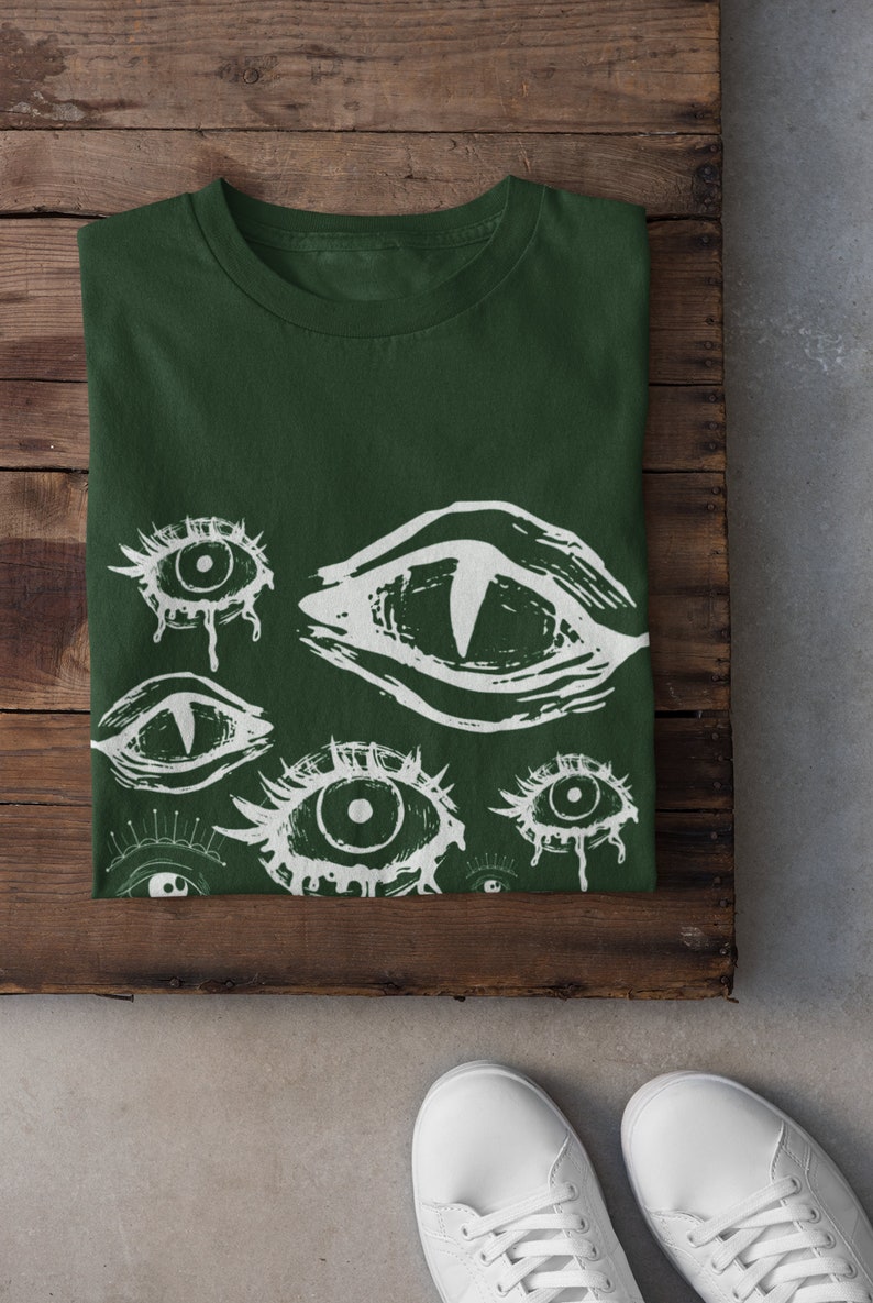 May include: Dark green t-shirt with a white eye graphic design. The design features multiple stylized eyes with various shapes and sizes. The shirt is folded on a wooden surface, with white sneakers visible.
