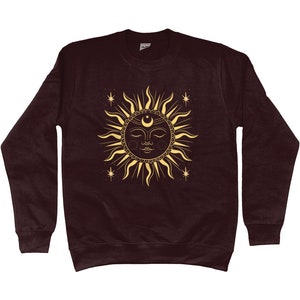 May include: A maroon sweatshirt with a gold sun and moon design. The sun has a face with closed eyes and a crescent moon above it. There are small stars around the sun.