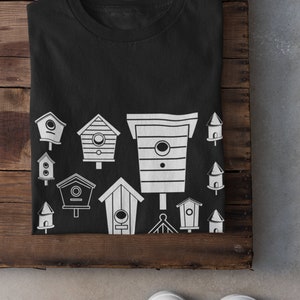 May include: Black t-shirt featuring a white design of various birdhouses. The birdhouses are of different shapes and sizes.