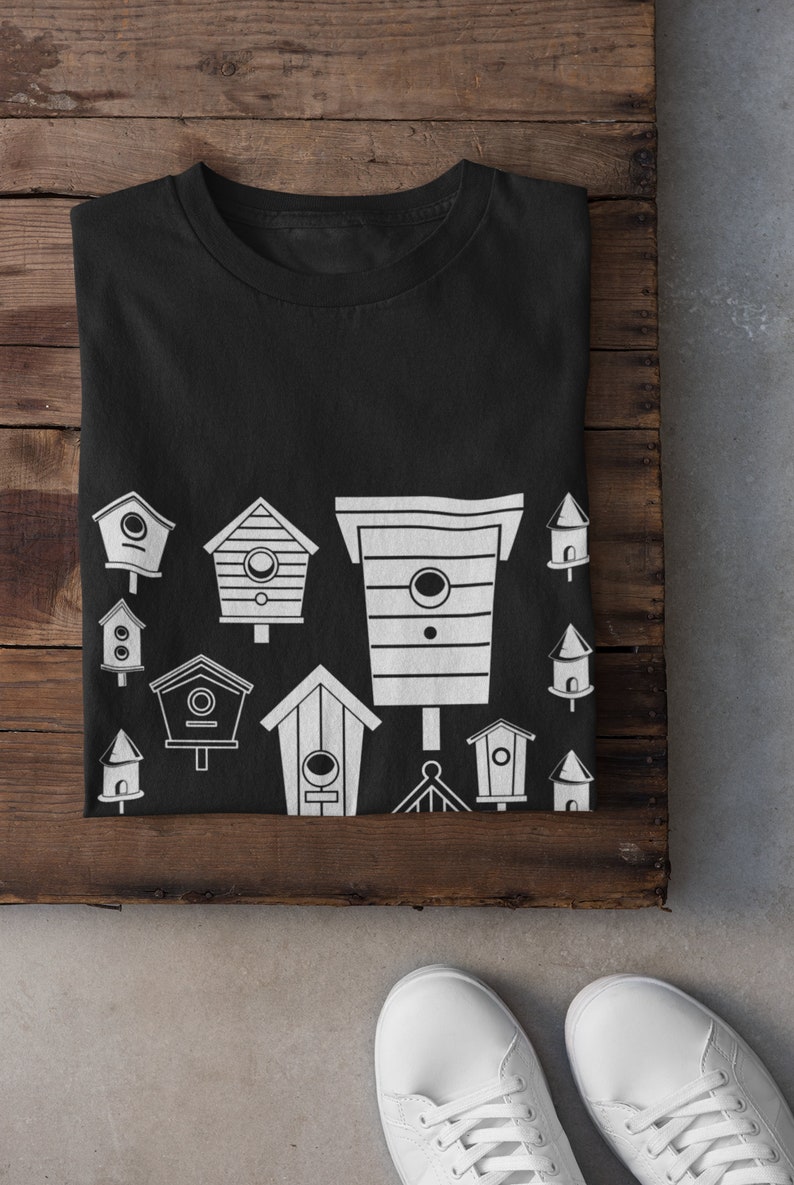 May include: Black t-shirt featuring a white design of various birdhouses. The birdhouses are of different shapes and sizes.
