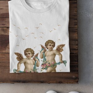 May include: White t-shirt with a print of two cherubs holding floral garlands and blue ribbons. The cherubs have wings and are surrounded by a pattern of flying birds.