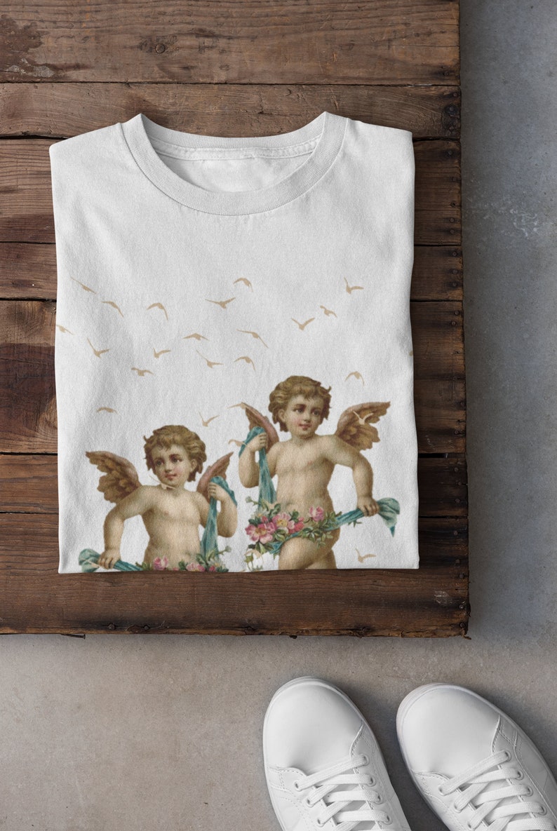 May include: White t-shirt with a print of two cherubs holding floral garlands and blue ribbons. The cherubs have wings and are surrounded by a pattern of flying birds.